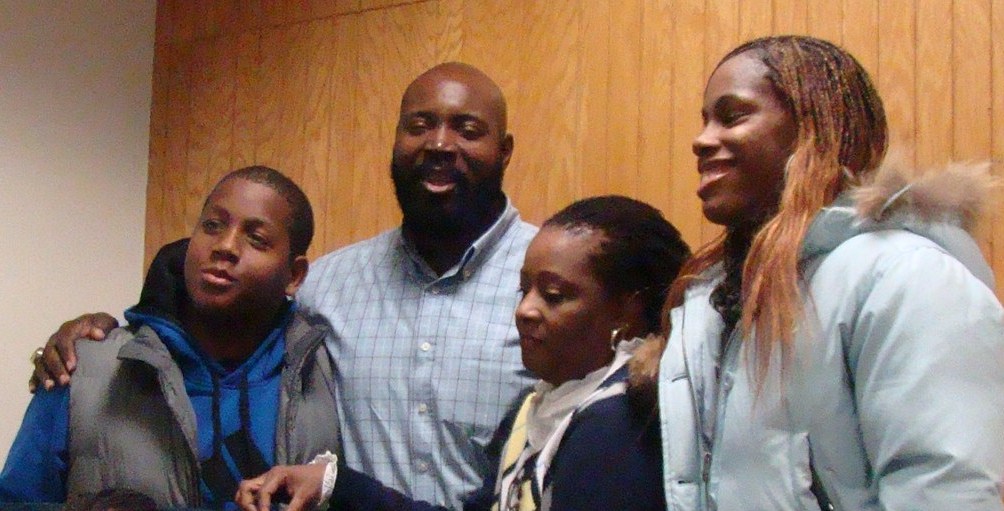 An adult man and woman, with their arms slung around 2 middle-high school aged kids.