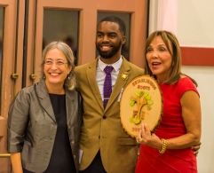 Two women and a man in the middle, all smartly dressed. The woman on the right in a red dress is holding up an oval plaque/award.