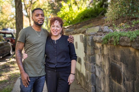 A young man and a woman posing together in front of a stone retention wall, a lush green backgdrop behind them.