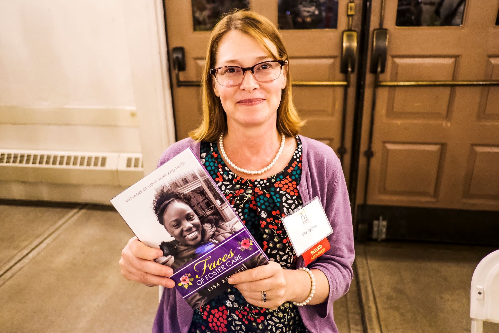 A blonde woman with glasses, holding a pamphlet. She has shoulder length blonde hair and is wearing a purple floral top with a nametag. Theresa Vargas.
