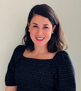Smiling woman with shoulder-length dark hair wearing a fitted black patterned dress against a plain light background.