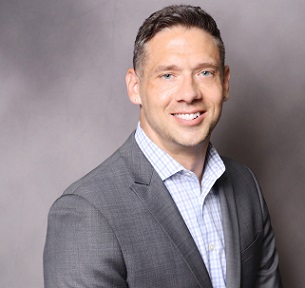 A confident, clean-cut professional with neatly trimmed dark hair and a warm smile, wearing a tailored gray suit jacket over a light checkered shirt against a soft gray backdrop.