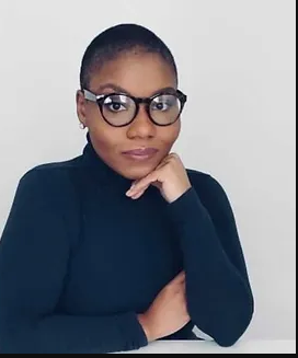 Woman with short hair and round glasses, wearing a dark turtleneck and resting her chin on her hand.