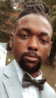 Man with neatly tied-back dreadlocks, wearing a light gray suit jacket and a patterned bow tie, looking thoughtfully at the camera.